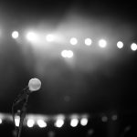 Microfone em um palco iluminado por fortes luzes, em uma imagem em preto e branco.