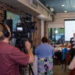 Videomaker registra um evento profissional em ambiente interno, com participantes e tela de projeção ao fundo.