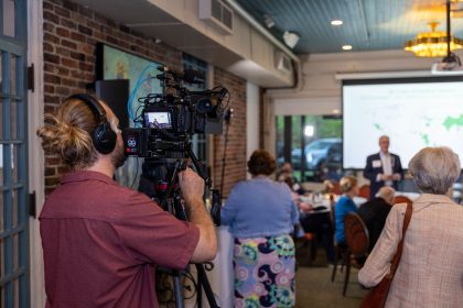Videomaker registra um evento profissional em ambiente interno, com participantes e tela de projeção ao fundo.