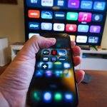 Mão segurando um celular com aplicativos de streaming diante de uma televisão