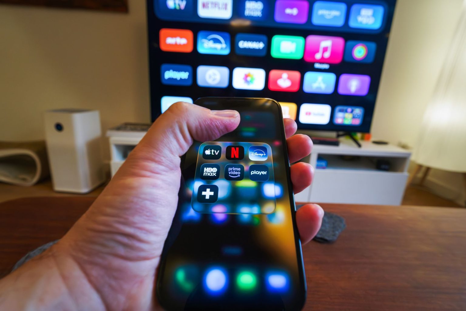 Mão segurando um celular com aplicativos de streaming diante de uma televisão