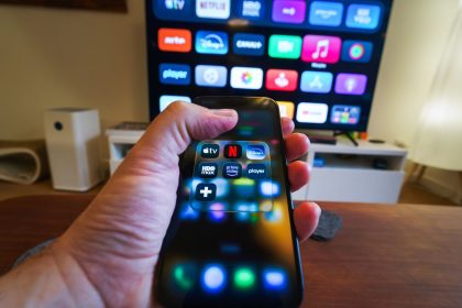 Mão segurando um celular com aplicativos de streaming diante de uma televisão