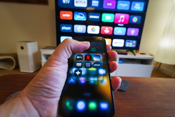 Mão segurando um celular com aplicativos de streaming diante de uma televisão