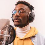 Cantor negro grava voz em estúdio usando fones de ouvido diante de um microfone