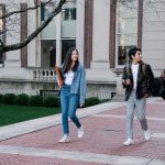Grupo diverso de estudantes caminhando por uma calçada do campus universitário em um dia ensolarado.