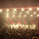 Banda de rock se apresenta no palco diante de um público, sob iluminação vermelha.