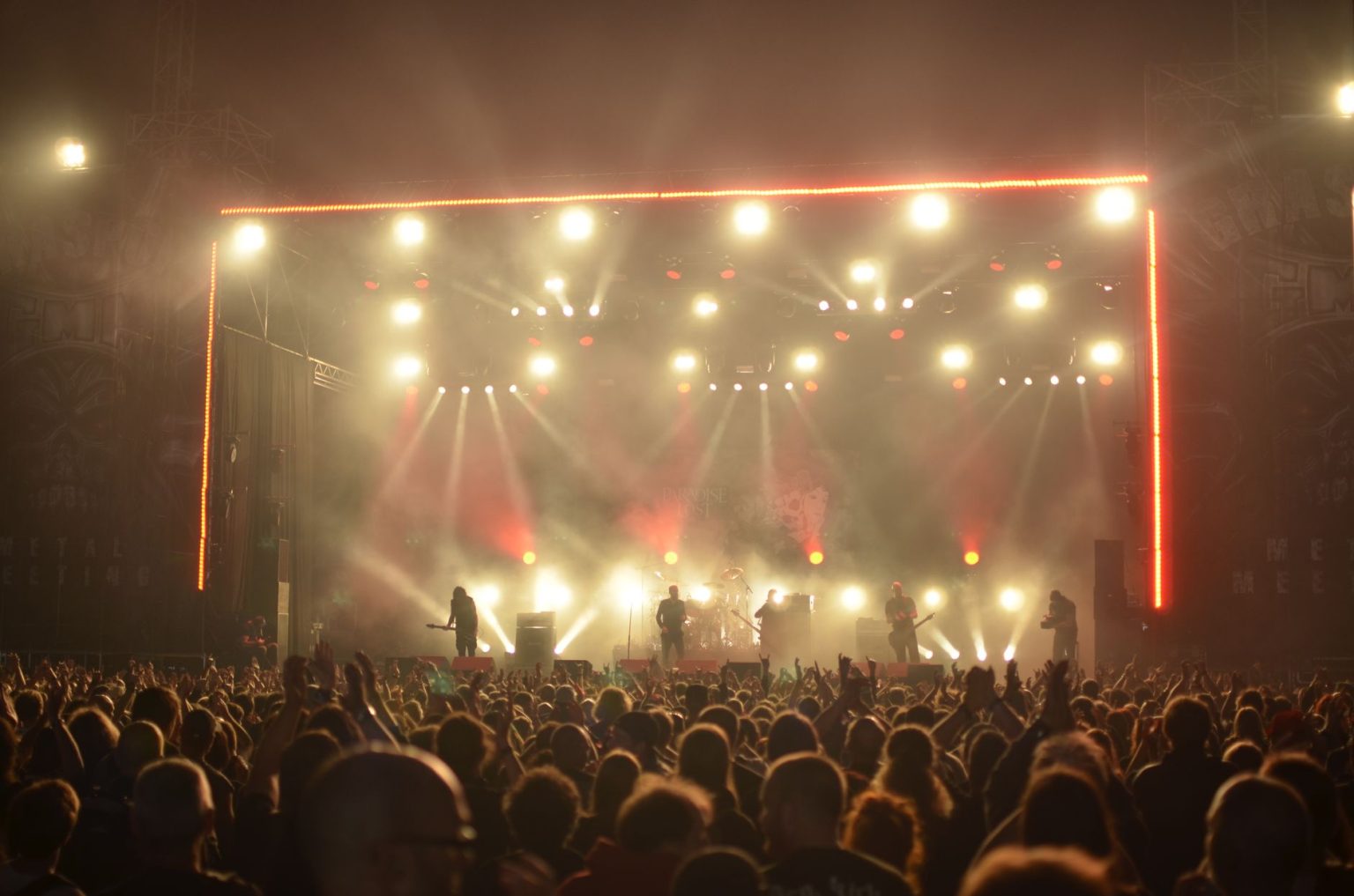 Banda de rock se apresenta no palco diante de um público, sob iluminação vermelha.