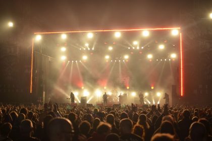 Banda de rock se apresenta no palco diante de um público, sob iluminação vermelha.