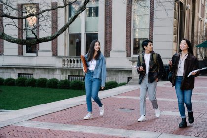 Grupo diverso de estudantes caminhando por uma calçada do campus universitário em um dia ensolarado.
