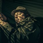 Homem observa pela persiana enquanto segura um telefone, em ambiente escuro e misterioso.