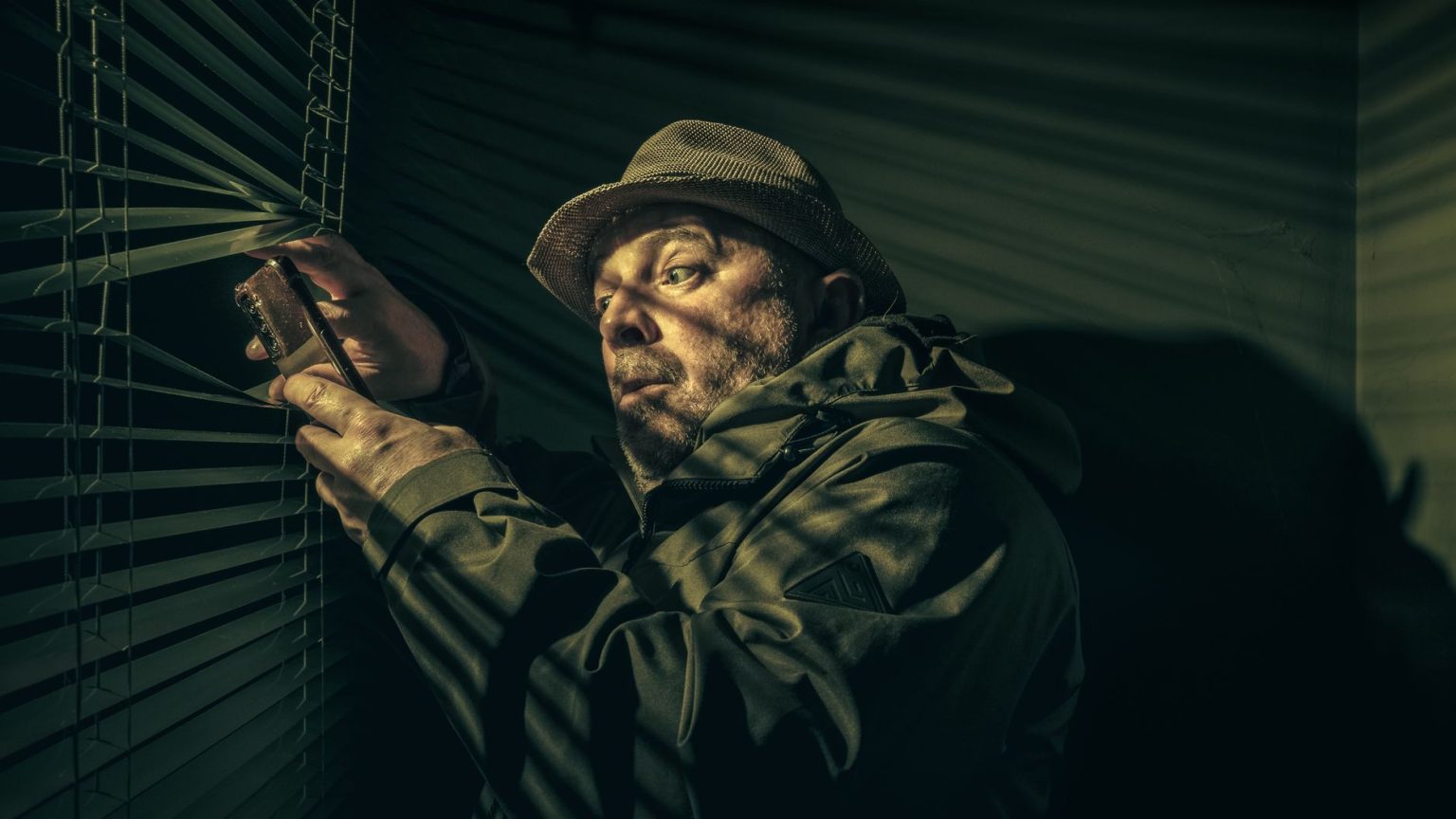 Homem observa pela persiana enquanto segura um telefone, em ambiente escuro e misterioso.