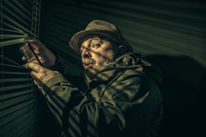Homem observa pela persiana enquanto segura um telefone, em ambiente escuro e misterioso.