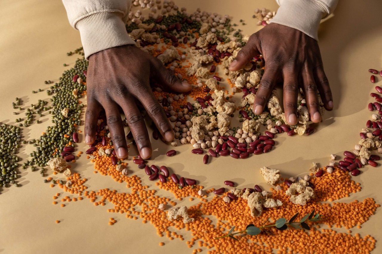 Mãos misturam grãos coloridos e ingredientes vegetais sobre uma mesa.