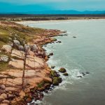 Vista aérea de trecho rochoso do litoral, com vegetação costeira e o mar ao fundo.
