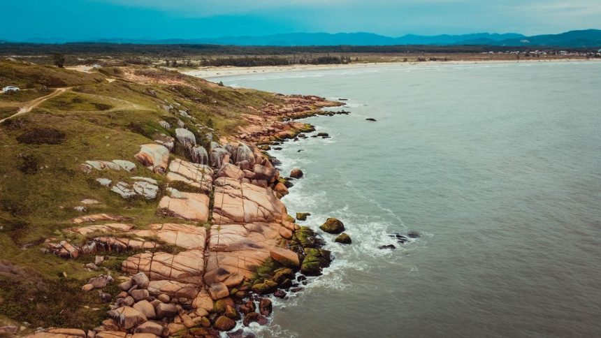 Vista aérea de trecho rochoso do litoral, com vegetação costeira e o mar ao fundo.