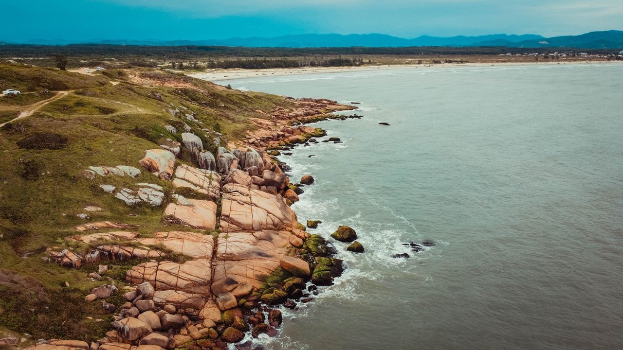Vista aérea de trecho rochoso do litoral, com vegetação costeira e o mar ao fundo.