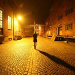 Mulher caminha sozinha por uma viela de paralelepípedos iluminada por postes de rua.