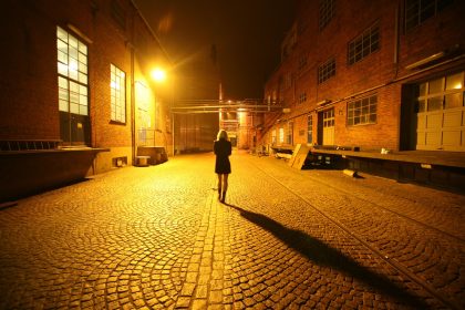 Mulher caminha sozinha por uma viela de paralelepípedos iluminada por postes de rua.