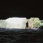Pessoa em silhueta na entrada de uma caverna, com a mata iluminada ao fundo.