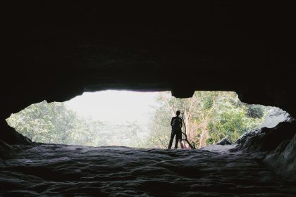 Pessoa em silhueta na entrada de uma caverna, com a mata iluminada ao fundo.