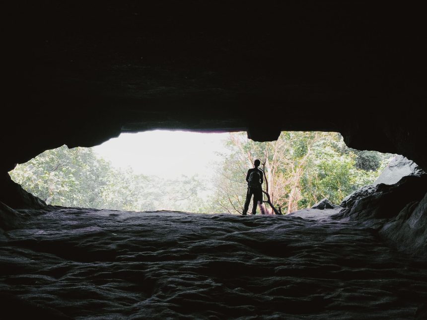 Pessoa em silhueta na entrada de uma caverna, com a mata iluminada ao fundo.